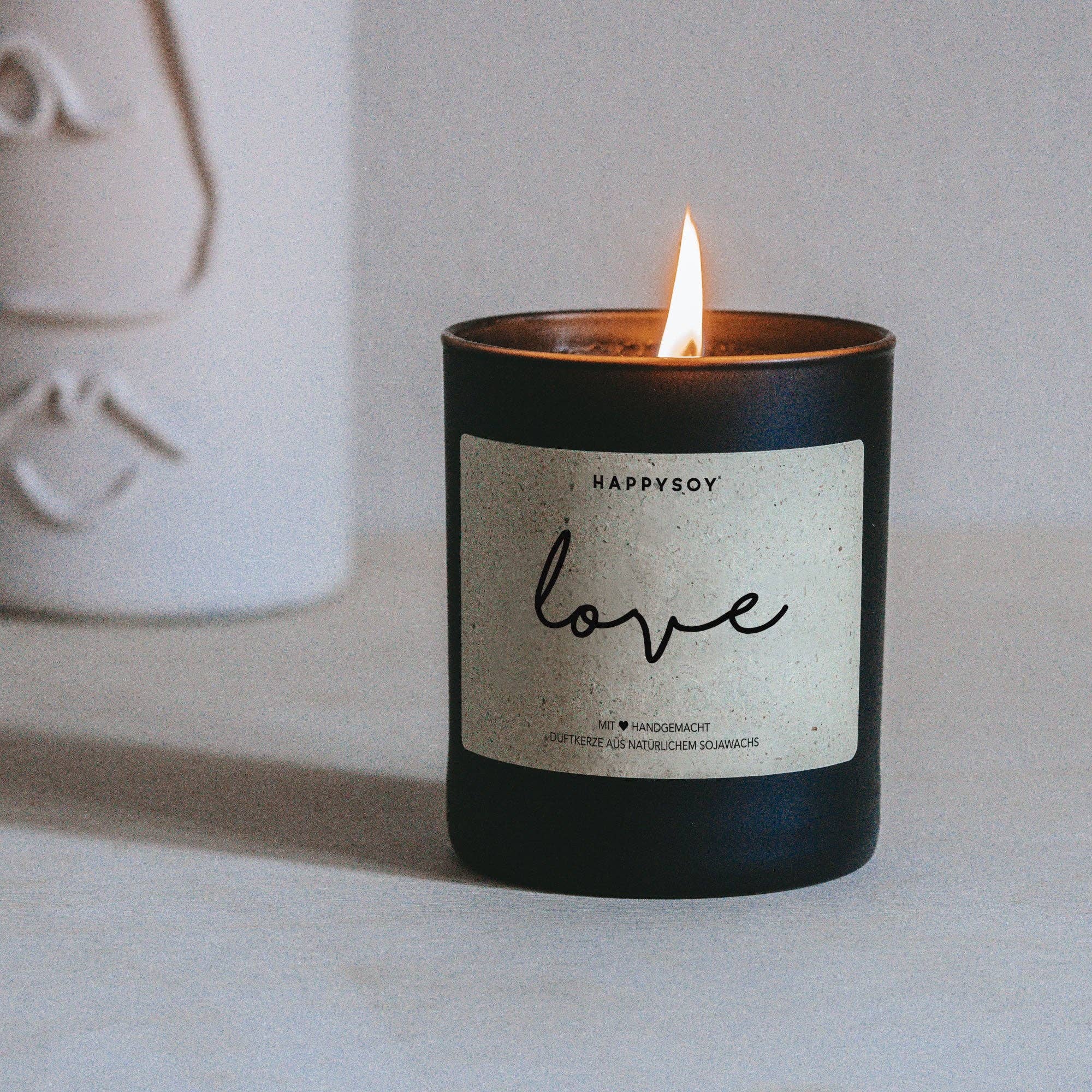 Love Duftkerze von Happysoy, Kerze in elegant Glas mit blumigem Duft, brennt auf einem weißen Tisch, warmes Licht.